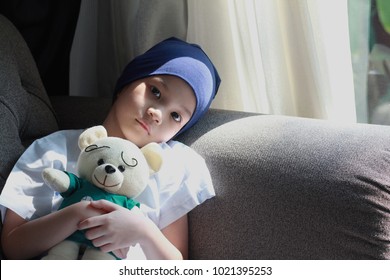 Patient kid lie down on couch or sofa in patient suit with her doll.Girl cover her head with  blue hat or headscarf.Kid look sad,tired and sick.Concept of   childhood cancer awareness.Selective focus.