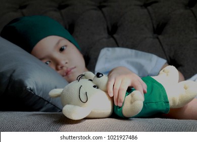 Patient Kid Lie Down On Couch Or Sofa In Patient Suit With Her Doll.Girl Cover Her Head With  Blue Hat Or Headscarf.Kid Look Sad,tired And Sick.Concept Of   Childhood Cancer Awareness.Selective Focus.