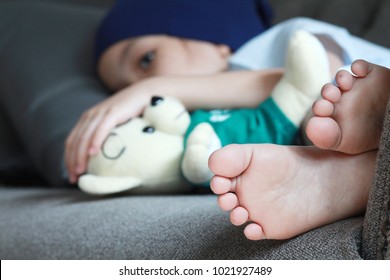 Patient Kid Lie Down On Couch Or Sofa In Patient Suit With Her Doll.Girl Cover Her Head With  Blue Hat Or Headscarf.Kid Look Sad,tired And Sick.Concept Of   Childhood Cancer Awareness.Selective Focus.