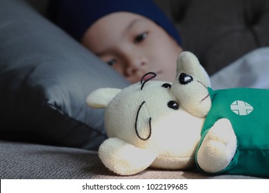 Patient Kid Lie Down On Couch Or Sofa In Patient Suit With Her Doll.Girl Cover Her Head With  Blue Hat Or Headscarf.Kid Look Sad,tired And Sick.Concept Of   Childhood Cancer Awareness.Selective Focus.