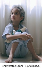 Patient Kid Sitting By The Window In Patient Suit With Her Doll.Skinny Little Asian Girl Look Sad,tired And Sick.Concept Of Childhood Cancer Awareness, Lonely Child Or Kid In Trouble Or Kid Violence.