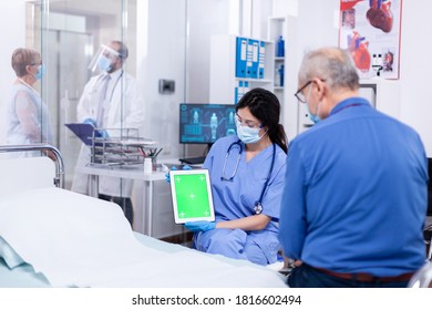 Patient Looking At Tablet With Green Screen During Consultation In Hospital Room With Nurse Wearing Face Mask Against Coronavirus As Safety Precaution. Medical Examination For Infections, Disease .