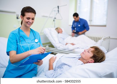 Patient Lying On Bed While Nurse Writing On Clipboard In Hospital