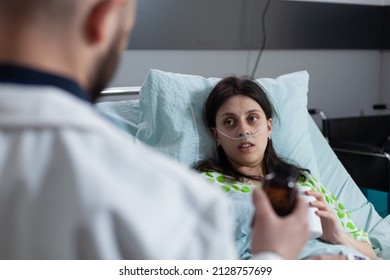 Patient With Nasal Cannula Recieving Oxygen Looking At Doctor Presenting Prescription Medication Bottle Recovering On Hospital Bed. Pharmacist Medic Giving Painkilers To Woman After Surgery.