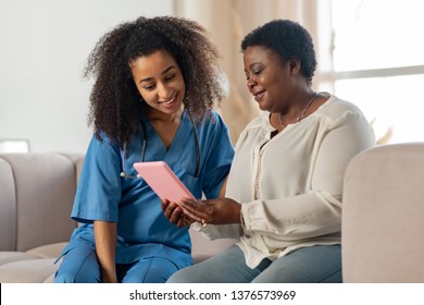 Patient And Nurse. Dark-skinned Pleasant Patient And Nurse Sitting On Sofa While Watching Movie On Tablet