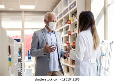 A Patient In A Pharmacy. Older Man With Gray Hair In An Elegant Suit Talks To A Woman Pharmacist And Explains Something With His Hands. Talk About Medical Therapy, Protective Mask Against Corona Virus