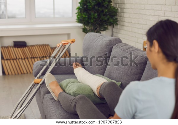 Patient with physical injury sitting on sofa at\
home with crutches beside. Woman with broken leg in plaster cast\
resting on couch in living-room. Rehabilitation and recovery after\
car accident concept