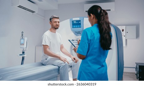 Patient pointing at form while speaking with nurse in turquoise scrubs holding clipboard. Discussing details of procedure before MRI session in bright diagnostic examination room. - Powered by Shutterstock - Get 15% off with code: PIKWIZARD15