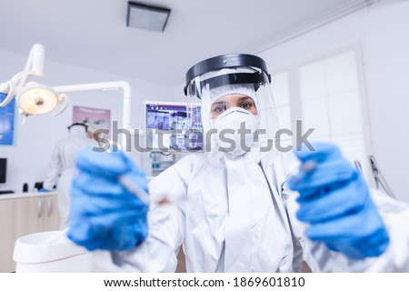 Patient pov of dentist examining patient gums using tools dressed up in covid suit in dental office. Stomatolog wearing safety gear against coronavirus during heatlhcare check of patient. [[stock_photo]] © 