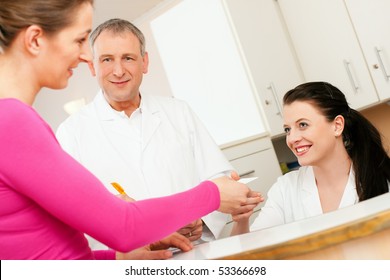 Patient in reception area of office of doctor or dentist, handing her health insurance card over the counter to the nurse, the doctor standing in the background and is writing things on a clipboard - Powered by Shutterstock