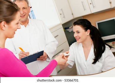 Patient In Reception Area Of Office Of Doctor Or Dentist, Handing Her Health Insurance Card Over The Counter To The Nurse, The Doctor Standing In The Background And Is Writing Things On A Clipboard