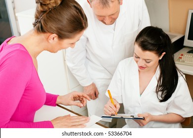 Patient In Reception Area Of Office Of Doctor Or Dentist, Handing Her Health Insurance Card Over The Counter To The Nurse Who Is Writing Things On A Clipboard, The Doctor Standing In The Background