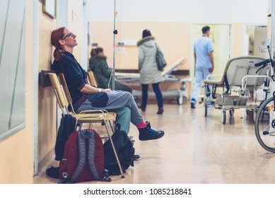 Patient Sitting In Hospital Ward Hallway Waiting Room With Iv. Woman With Intravenous Therapy In Her Hand Is Waiting In The Clinic Corridor With Blurred Medical Personnel In Background.