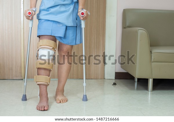 Patient\
standing on crutch in hospital ward ware knee brace support after\
do posterior cruciate ligament surgery ,Bandage on knee of asian\
woman on crutches.healthcare and medical\
concept.