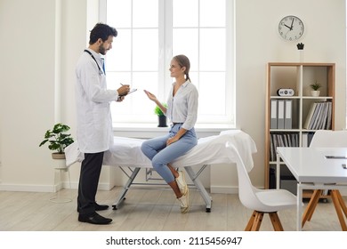 Patient Talking To A Doctor At A Modern Clinic Or Hospital. Male Physician In A White Coat Giving A Consultation To A Young Woman Who Is Sitting On The Medical Couch In The Exam Room