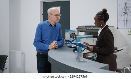 Patient talking to female receptionist before attending appointment with doctor at medical facility center. Woman helping old man with checkup report papers at hospital reception desk. - Powered by Shutterstock