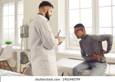 Patient Telling Physician About His Pain And Health Problems During Visit To Hospital. Young African-American Man Complaining About Back Ache While Sitting On Examination Bed At The Doctor's Office