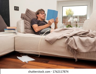 Patient with urethral catheter inserted into the penis, reads a book lying on the sofa at home.