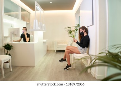 Patient waiting at the reception of the dental, gynecological or aesthetic clinic. The patient is using her smartphone while the receptionist takes a call. Medical concept.