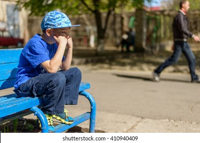 Patient young boy sitting waiting on a blue wooden outdoor bench in a trendy blue outfit with his hand to his chin and a bored expression - Powered by Shutterstock