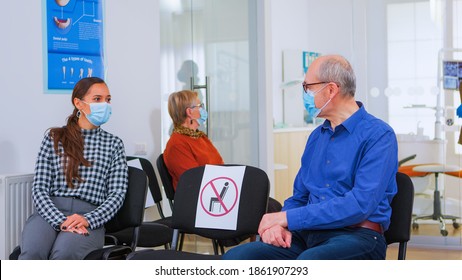 Patients With Protection Mask Talking Sitting On Chairs Keeping Social Distance In Stomatological Clinic, Waiting For Doctor During Coronavirus. Concept Of New Normal Dentist Visit In Covid19 Outbreak