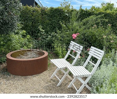 Patio with blooming roses, pair of white antique folding chairs and Corten steel fountain