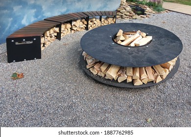 Patio Fire Pit Round Table With Log Holder On The Stone Or Gravel Surface. Iron Rounded Fire Pit With Grill Top On Backyard Party Place. Grill Appliance And Fireplace On The Backyard. Iron Furniture.