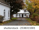 Patrimonial white wooden house with silver metal sheet steep roof seen at the end of an alley during a fall morning, with side of other old building in the foreground, Beaumont, Quebec, Canada