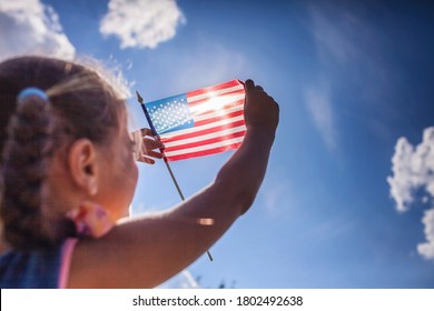 Patriot and National Flag day celebration. Cute little patriot sitting on the meadow and holding the national flag of United States over blue sky background, summer outdoor, soft focus on flag