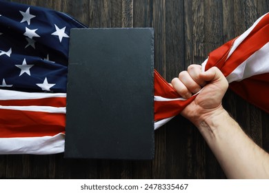 Patriotic Display of Strength: Hand Gripping American Flag in front of Blank Slate on Wooden Background - Powered by Shutterstock