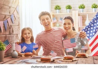 Patriotic holiday. Mother, father and daughter are doing burgers. Happy family are celebrate 4th of July. Cute little child girl with American flag in the room at home.