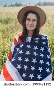 Patriotic Portrait Of Beautiful Young Woman In Beige Hat Wrapped In American Flag Standing On Meadow At Sunny Day. Hipster Girl Smiling And Looking At Camera. July 4th Forth Independence Day Concept