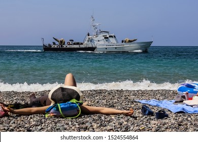 A Patrol Boat On The Horizon (coast Guard), And A Sunbathing Girl In A Hat On A Blurred Foreground