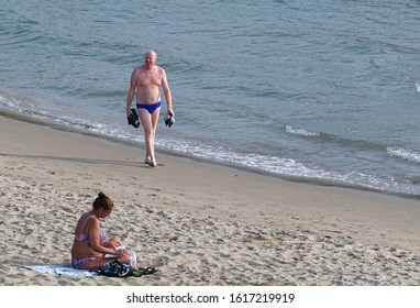 Pattaya, Thailand - December 23, 2019: Bald Older Man Walking On The Beach. Woman Sits And Reading A Book.