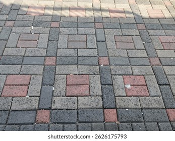 A patterned sidewalk made of interlocking paving blocks in shades of red and gray, forming a repetitive geometric design across the surface.