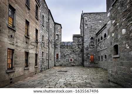 Paved courtyard of ancient prison, surrounded by massive stone walls with lit windows. Kilmainham Gaol, Dublin, Ireland.