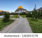 Paved foot path cuts through pine trees as it leads to a sunlit mountain peak at dawn near Logan Pass in Glacier National Park, Montana. 