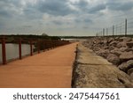 A paved path with a crimson railing runs by a rugged embankment under an overcast sky. It leads over a dam, offering water views and a scenic landscape in Brushy Creek Lake Park, Cedar Park, Texas.