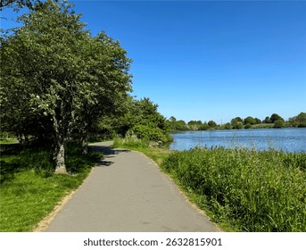 A paved path curves alongside a tranquil tarn, bordered by lush green grass and leafy trees. The blue sky and sunlight enhance the peaceful atmosphere over, Yeadon Tarn, Yeadon, Leeds, UK