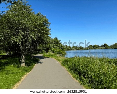 A paved path curves alongside a tranquil tarn, bordered by lush green grass and leafy trees. The blue sky and sunlight enhance the peaceful atmosphere over, Yeadon Tarn, Yeadon, Leeds, UK
