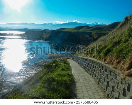 Paved path winds along scenic, grassy cliffside overlooking sparkling Pacific ocean along Kaikoura Coast Track, Canterbury, New Zealand. Rolling hills, snow-capped mountains in view. Coastal trails