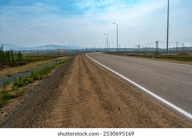 A paved road with a dirt shoulder curves through a rural landscape, surrounded by vegetation and distant mountains under a cloudy sky. Power lines stretch into the distance, with no people in sight - Powered by Shutterstock