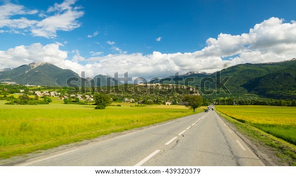 Paved two lane road crossing mountains and forest\
in scenic alpine landscape and moody sky. Panoramic view from car\
mounted camera. Summer adventure and roadtrip in the Italian French\
Alps.