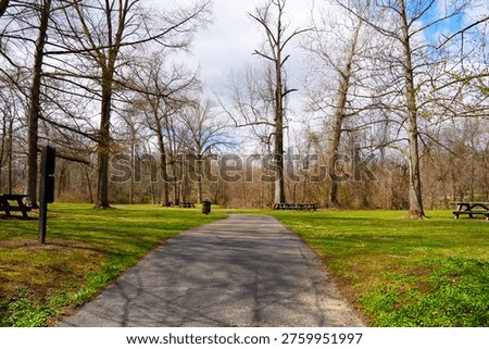 A paved walking and biking path winds through the scenic Newport Wetlands in Boardman, Ohio, during the transition from late winter to early spring. This well-maintained area within Mill Creek MetroParks features numerous picnic tables shaded by tall, bare deciduous trees under a cloudy blue sky. The vibrant green grass of the recreation area provides a natural contrast, creating a peaceful and inviting atmosphere. Ideal for local tourism guides, park management materials, health and wellness advertisements, and regional geography illustrations of Mahoning County and the Midwestern United States.
