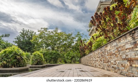 Paved Walking Path In A Tropical Park. There Is A Stone Fence On The Side. Nearby, In An Ornamental Pool, Lush Green Vegetation. Clouds In The Sky. Seychelles.  