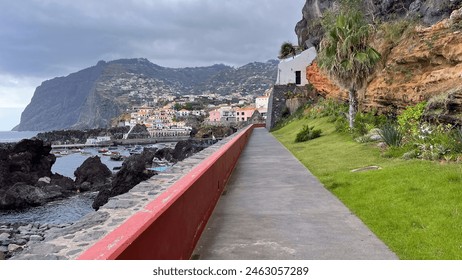 A paved walkway runs along the rocky coastline with a view of a cliffside town, featuring colorful houses and a mountain in the background under an overcast sky - Powered by Shutterstock