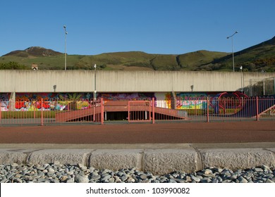 A Pavement In Front Of A Fenced Skate Park With Ramps And A Graffiti Wall Behind With Hills And A Clear Blue Sky In The Distance.