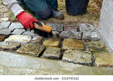 Pavement Works, Cobbled Pavement Natural Stone. Gloved Hands Of Worker Cleaning Concrete Paver Blocks With Brush And Water After Laying Paving Stones Carefully Placing In Cement, Building A Ramp Way