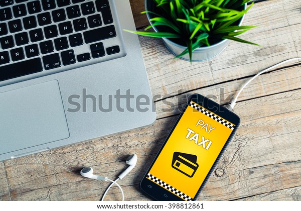 Pay Taxi message on a mobile\
phone screen. Office wooden desk with laptop computer and a\
plant.