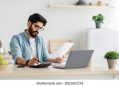 Paying bills, taxes at home online. Young arab man using calculator and laptop computer, calculating taxes or planning budget while sitting at kitchen table at home, copy space
